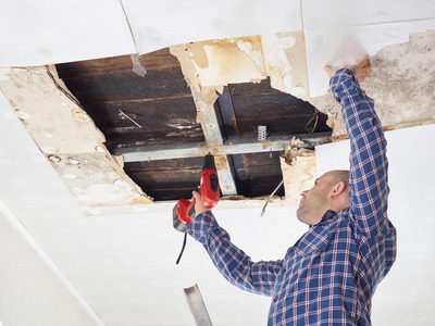 guy with a blue shirt repairing the inside portion of a roof on a ladder with power tools