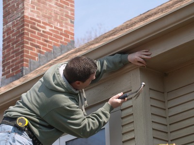 man in a green jacket and a hammer appearing to be repairing the top of a home